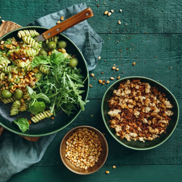 Summer vegetarian pasta salad with broccoli pesto, peas, arugula, olives, pine nuts and bread crumbs on dark green background. Top view.